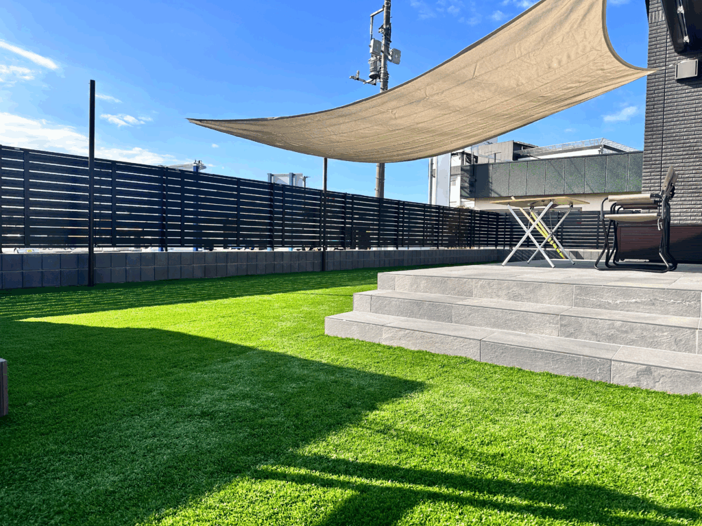 M様邸の新築外構は、 ブラックとグレーをベースにした統一感のあるデザインが特徴です。 木目調の部分も、あえて暗めの色を選び、 建物とよくなじむ落ち着いた印象に仕上げました。 お庭には、35mmのふかふか人工芝を使用。 ワンちゃんが走っても足にやさしく、 広々としたドッグランとして楽しめます。 門扉やフェンスは、角を丸くした形を選び、 すき間や高さにも気を配りました。 ブロックを含めて約1.6mの目隠しフェンスで、 外からの視線もしっかりカットしています。 また、 上げ下げできるシェード 人感センサー付きライト 落とし棒を2本付けた動きにくい門扉 など、 ワンちゃんもご家族も安心できる工夫をたくさん取り入れました。